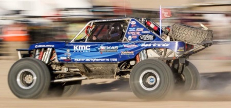 A photo of a blue ultra 4 style racecar driving fast through the desert